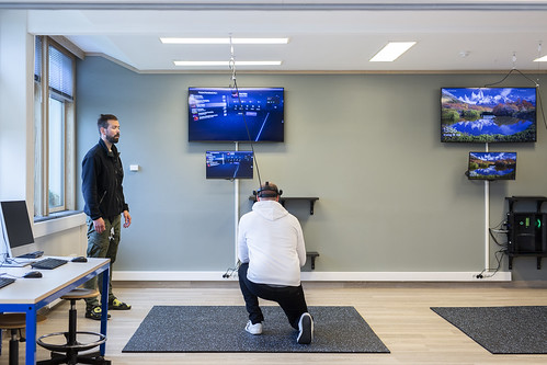 En person som står ved siden av en person som sitter på en stol i et rom med flere skjermer