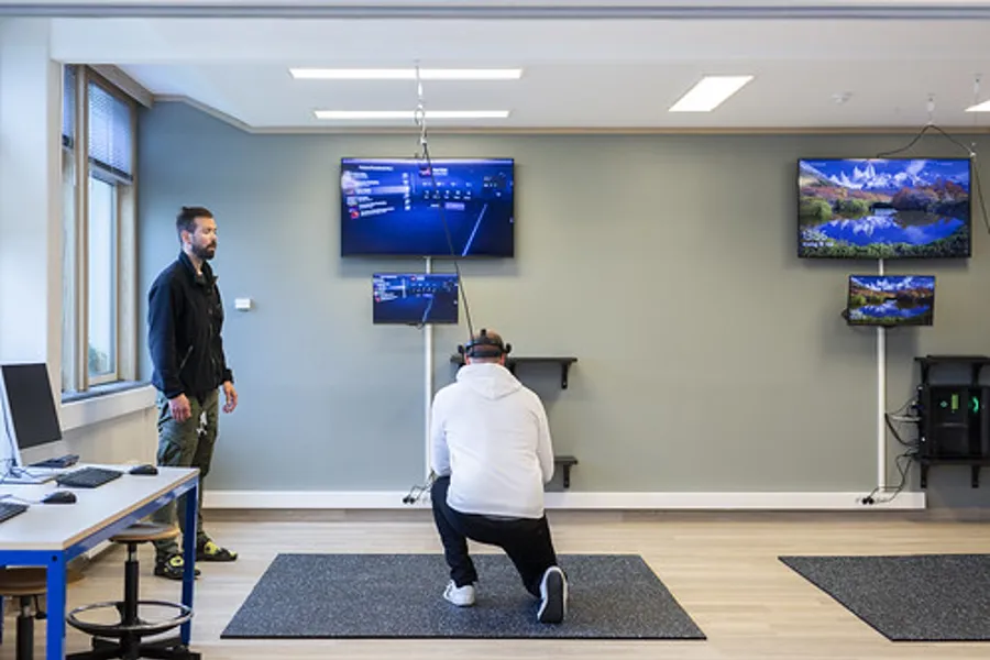 En person som står ved siden av en person som sitter på en stol i et rom med flere skjermer