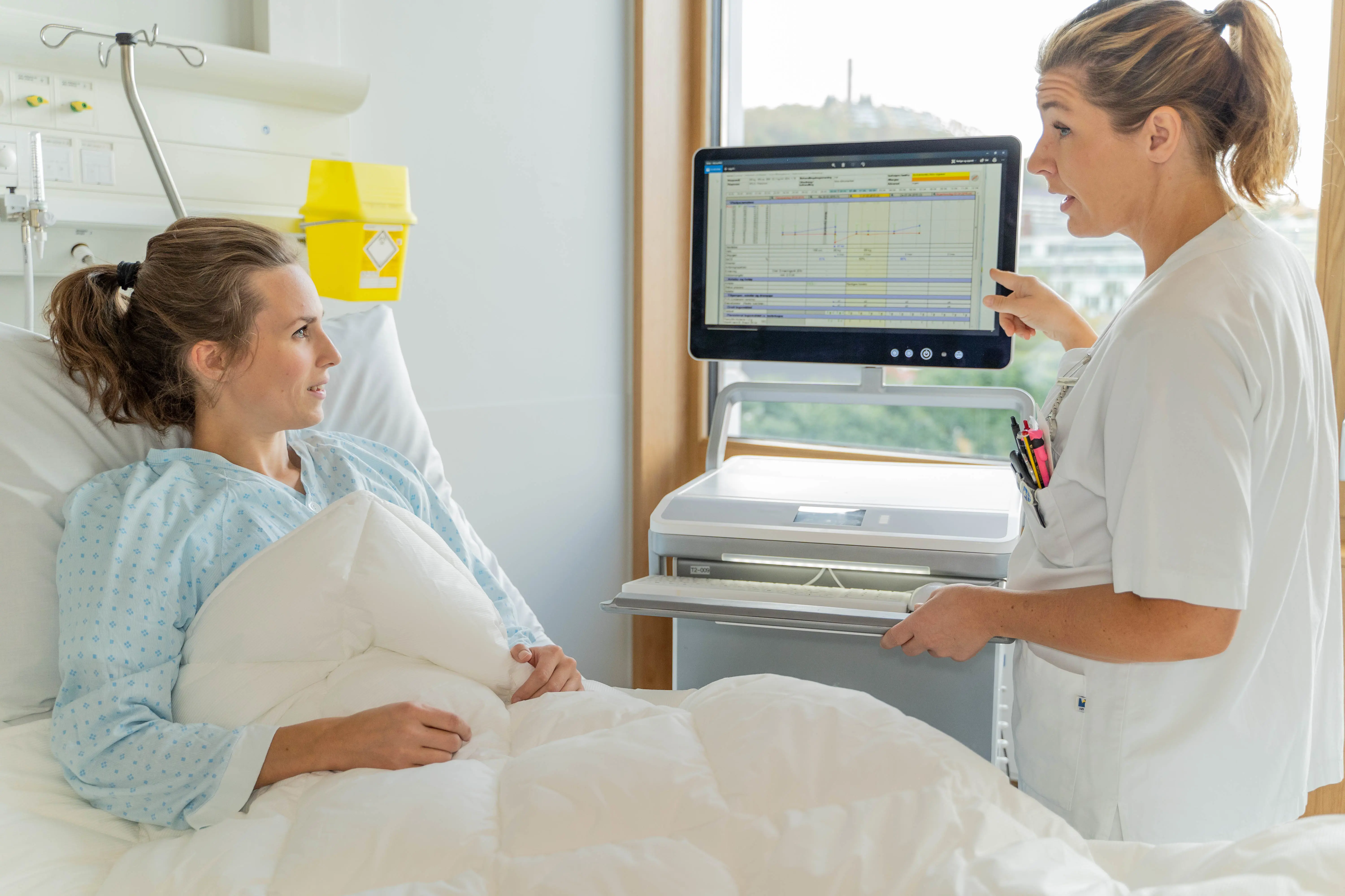 Pasient i senga, skepleier og legemiddeltralle. Foto.