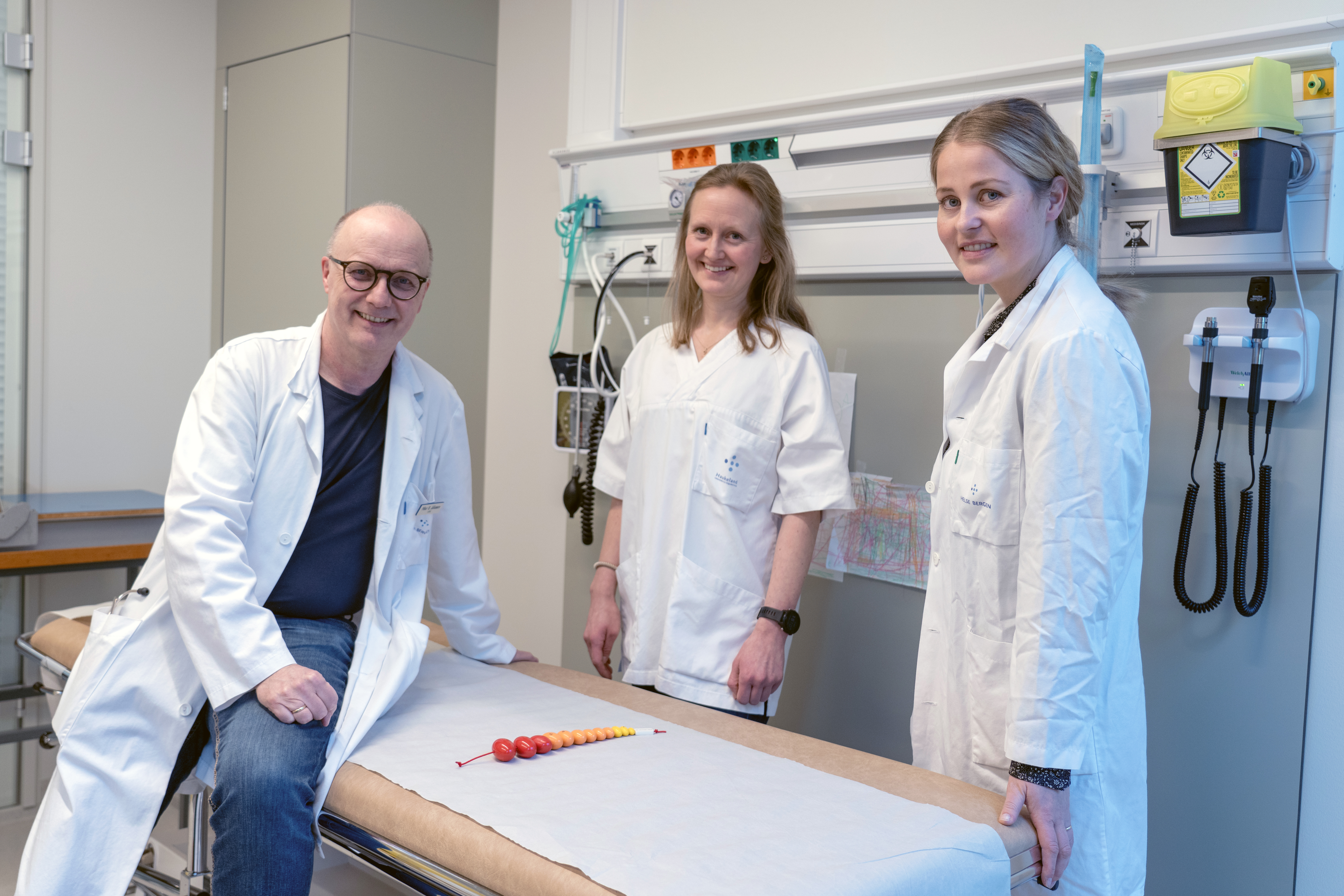 En mann og to kvinner i hvite frakker ved et undersøkelsesbord.