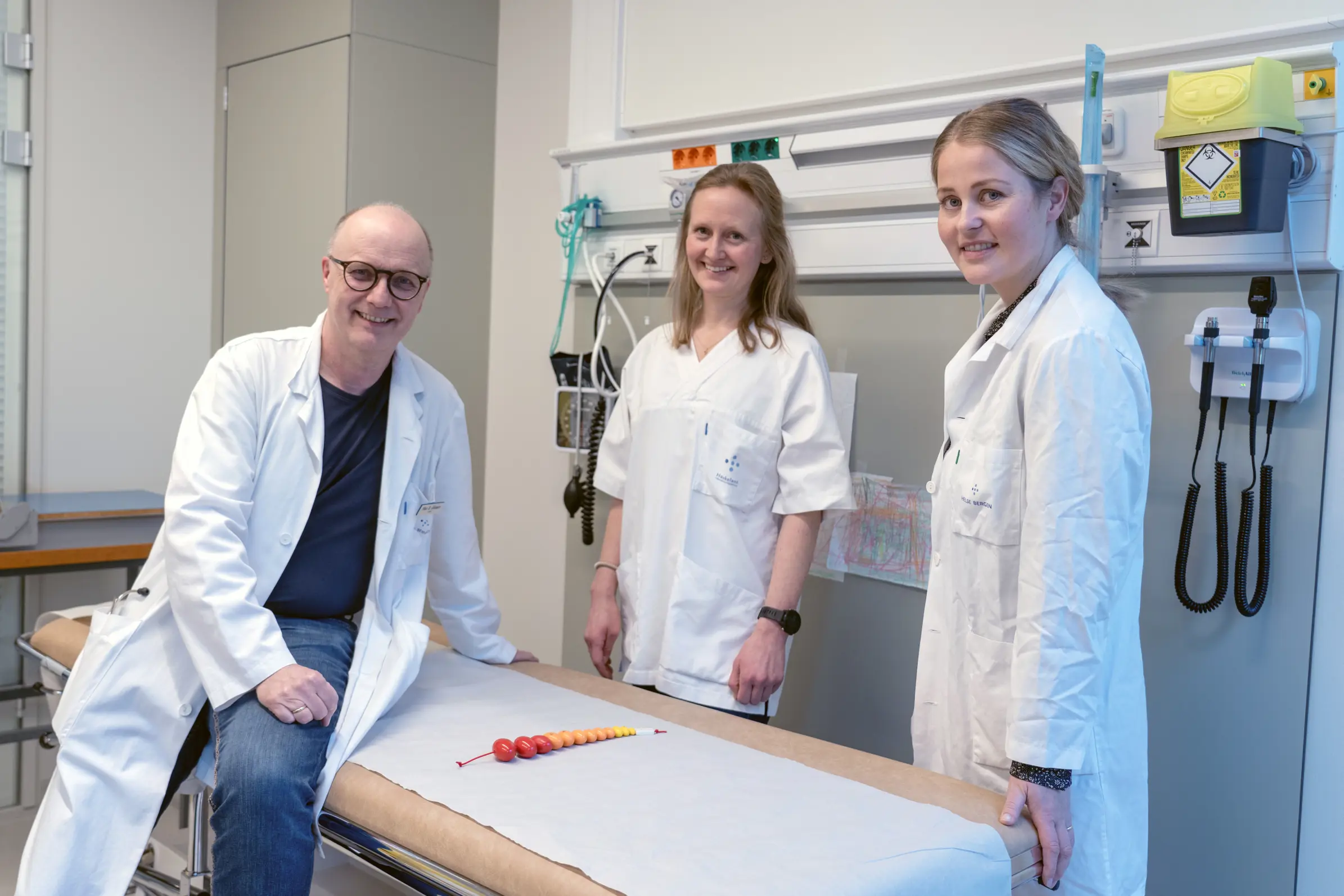 En mann og to kvinner i hvite frakker ved et undersøkelsesbord.