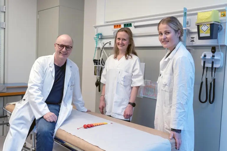 En mann og to kvinner i hvite frakker ved et undersøkelsesbord.